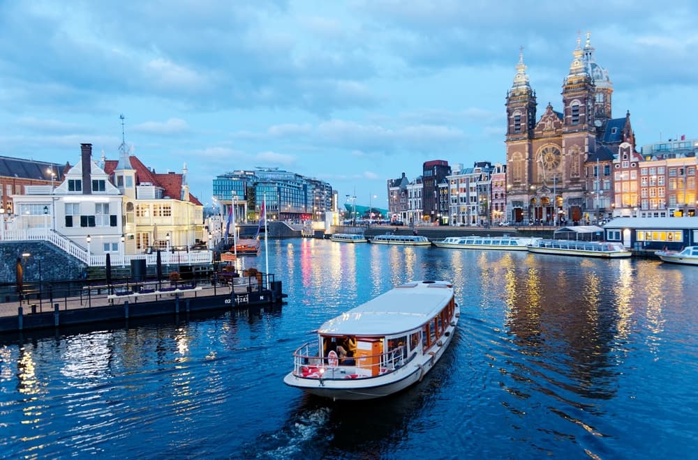 A tour boat cruising on the canal near Amsterdam's Central Railway Station, with the spires of Saint Nicholas Basilica towering in background and city lights reflected in the water at blue dusk