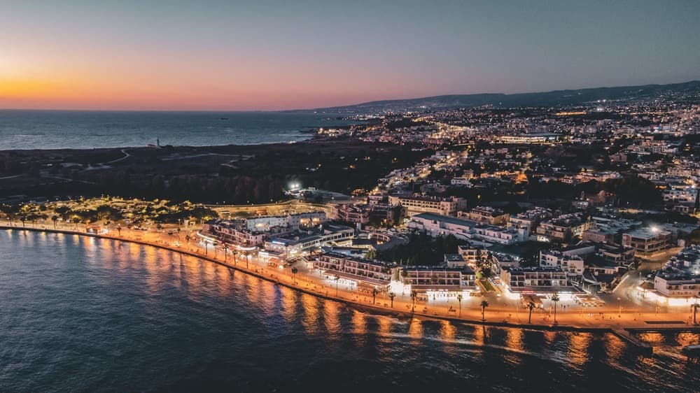 Aerial view of Kato Paphos Harbour, Cyprus at dusk, with illuminated buildings and a calm sea reflecting the vibrant sunset.